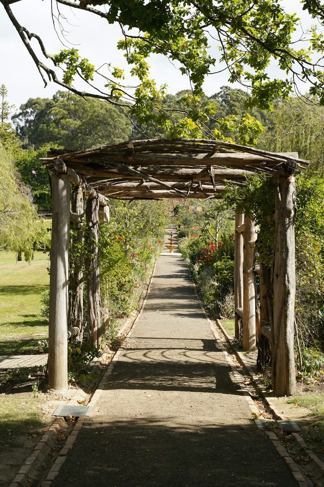 A garden path leads through a wooden pergola toward a fountain.